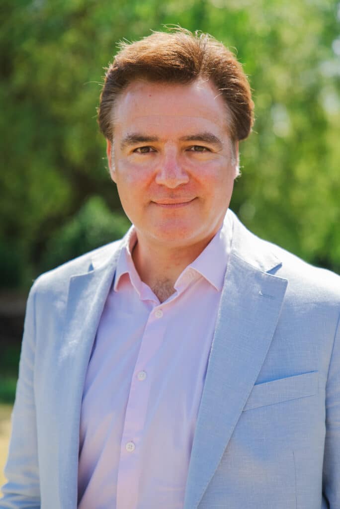 A chest-up portrait of Andrew Whelan, Founder and CEO of Westcott Care, looking directly at the camera with a subtle smile. He is dressed in a light blue linen blazer over a pink shirt, set against a soft-focus background of lush green trees on a sunny day.