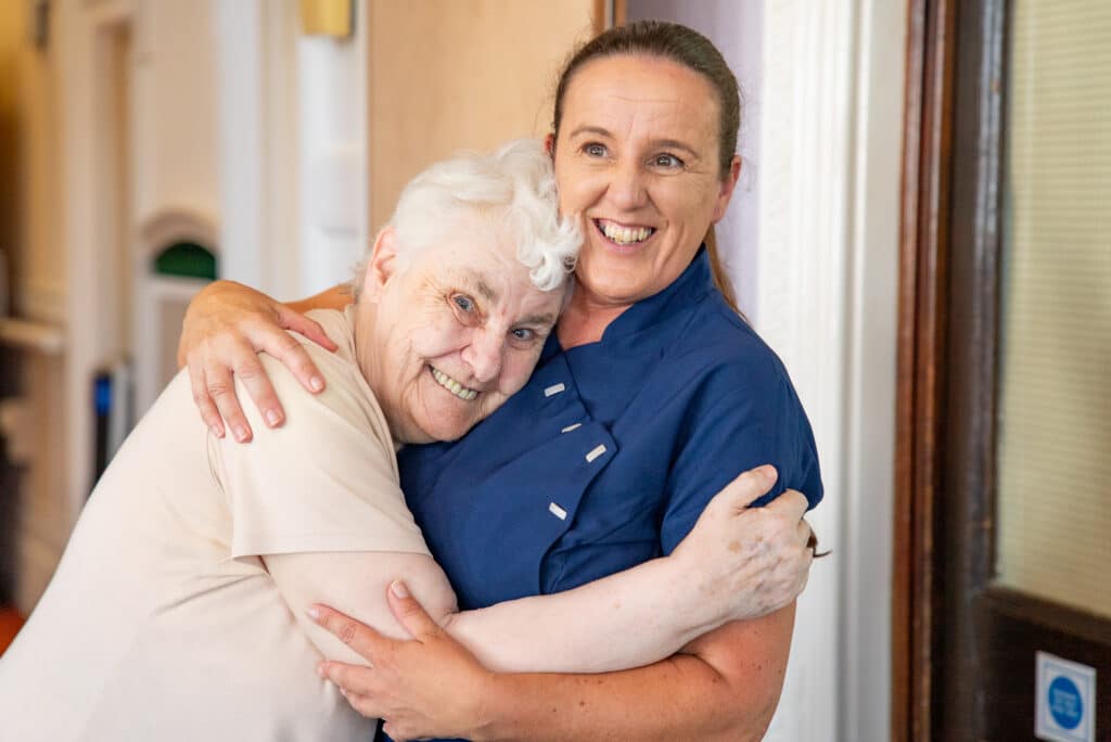 A heartwarming, candid moment in a carpeted hallway at Cartmel Grange care home. A female carer in a navy tunic and a senior female resident wearing a light beige t-shirt are embraced in a tight, affectionate hug, both smiling with happiness and comfort.