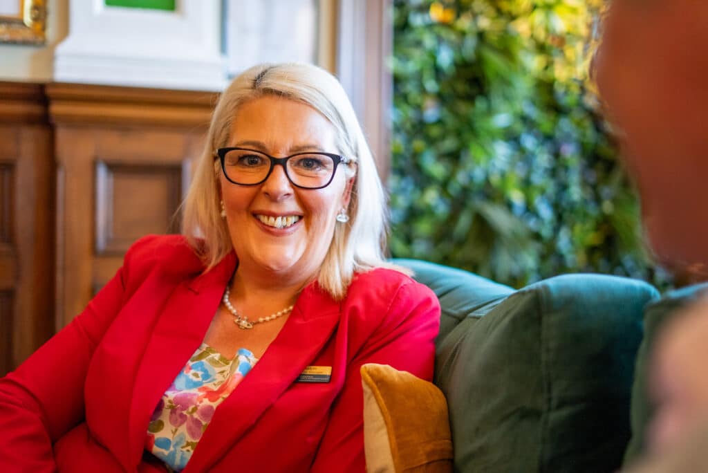 A warm indoor portrait of Registered Manager Shelly Ralph at Cartmel Grange care home. She has straight blonde hair, wears black-rimmed eyeglasses, a pearl necklace, a floral patterned top, and a vibrant red blazer with her staff name badge. She sits comfortably on a plush green armchair, smiling broadly during a conversation.