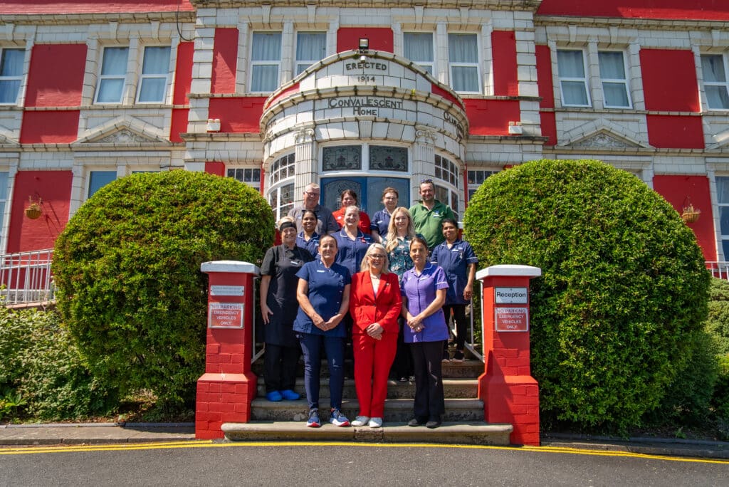 A group photograph of eleven care home staff members standing together on the stone entrance steps outside Cartmel Grange care home. A woman in a bright red suit stands front and center, flanked by staff in various blue and purple uniforms. The grand building in the background features vibrant red painted walls with white stone trim and historic architectural text reading "ERECTED 1914 CONVALESCENT HOME" above the arched doorway, framed by two large rounded green bushes.