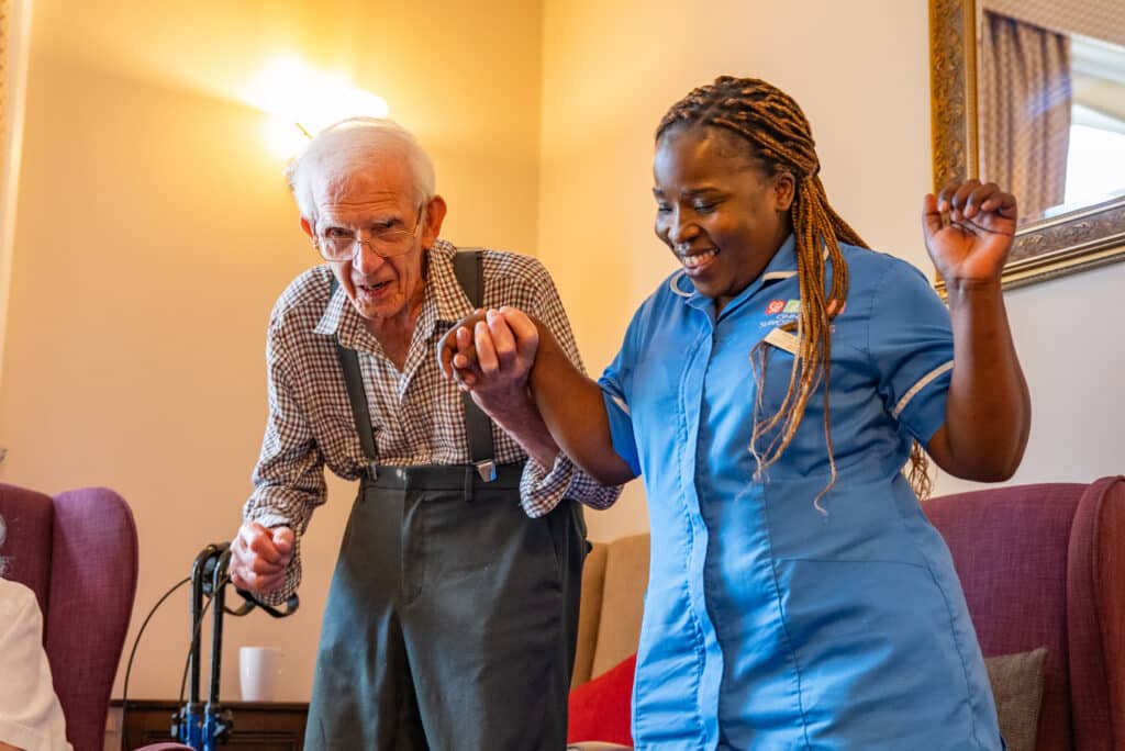 A female care assistant wearing a blue uniform laughs joyfully while holding hands and dancing with an elderly male resident wearing glasses and braces, illustrating interactive care activities and physical wellbeing for seniors.