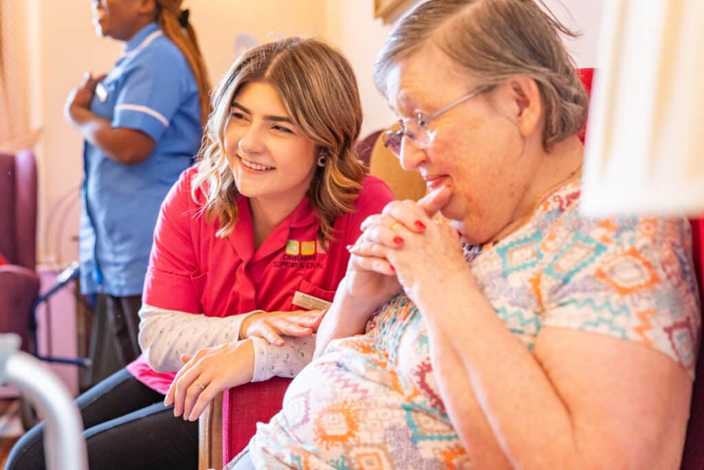 A smiling young female care assistant in a bright pink uniform leans in to share a warm, compassionate moment with an elderly resident, showcasing the dedicated person-centred care and companionship at Doddlespool Hall.