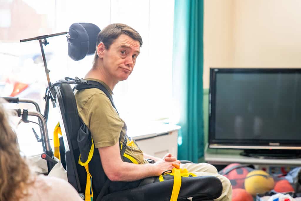 A man sitting comfortably in a specialized, fully supported wheelchair inside a bright lounge area at Linford Grange, looking toward the camera during an indoor group activity.