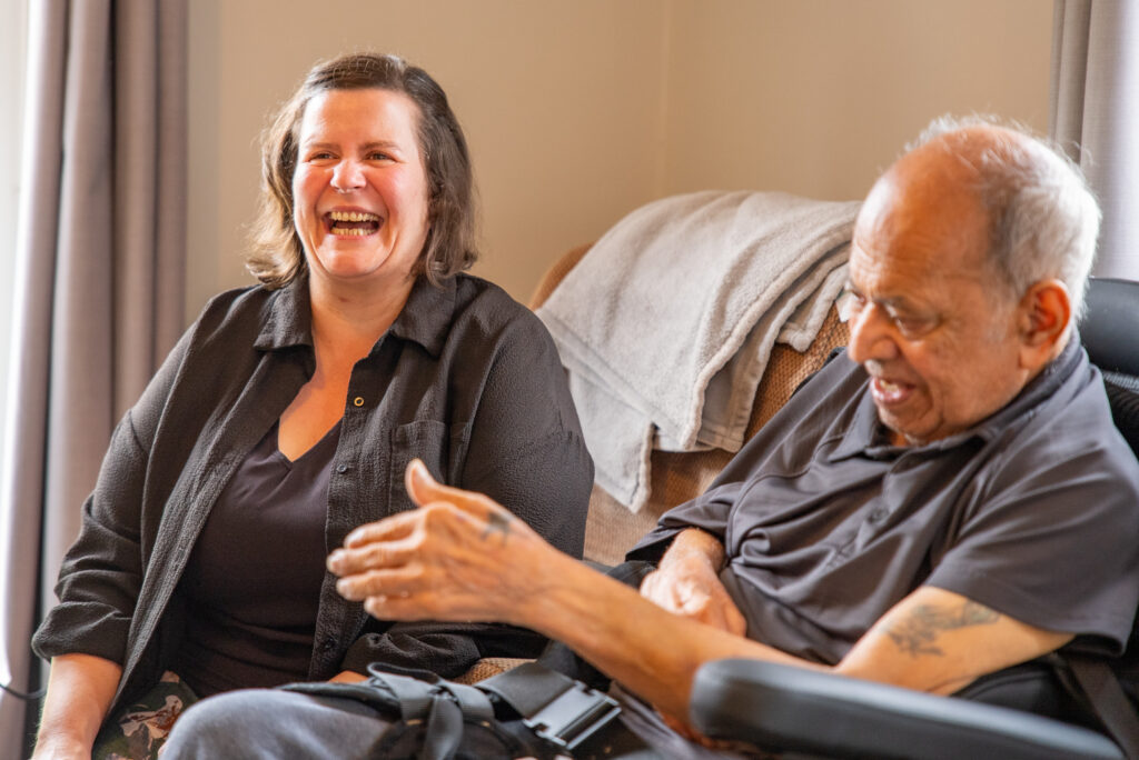 Medium close-up of a woman in a dark shirt laughing freely while sitting next to an elderly man in a black wheelchair who is smiling and gesturing with his hands.
