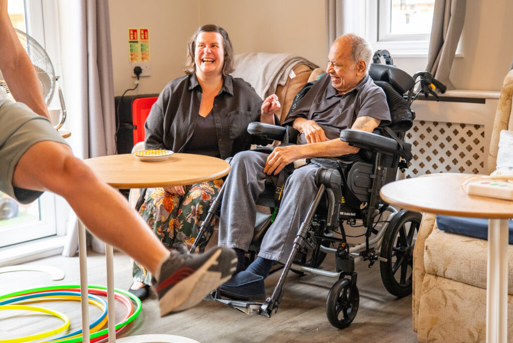 An elderly man in a black wheelchair and a woman seated next to him laugh during an indoor exercise activity at a care home. A person's leg extended in motion is visible in the foreground near colorful hula hoops scattered on the floor, while a small wooden table sits between them in a brightly lit, comfortable room.